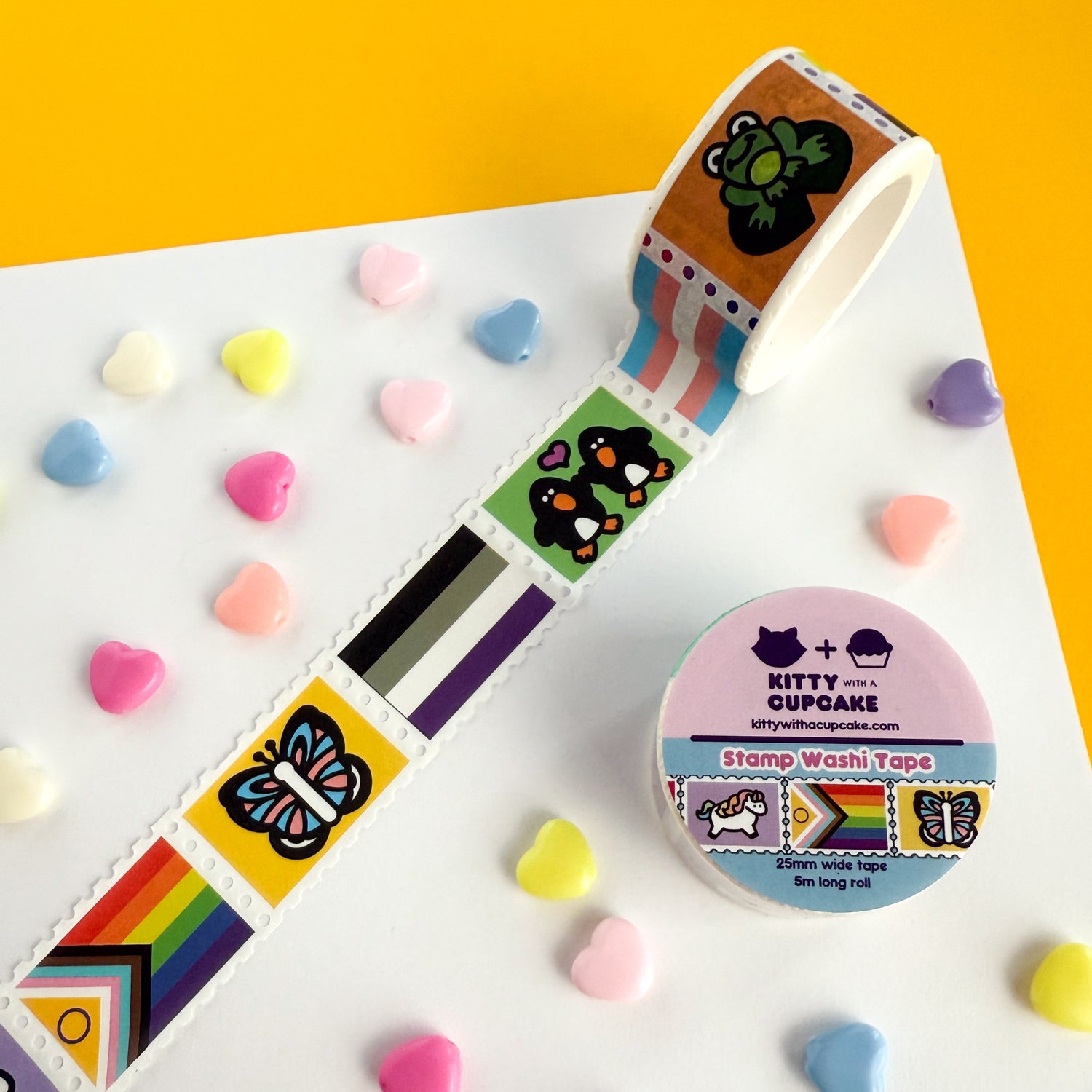 A roll of pride themed stamp washi tape. There is a packaged roll of tape with the Kitty With a Cupcake logo and an example of the tape inside. Next to that is a displayed section of the tape, which is sectioned into stamp-style squares, and around the two are decorative heart beads.