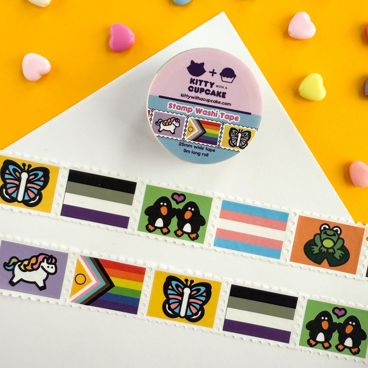 A roll of pride themed stamp washi tape. There is a packaged roll of tape with the Kitty With a Cupcake logo and an example of the tape inside. Below are two displayed section of the tape, which is sectioned into stamp-style squares, and above are decorative heart beads on a yellow background.