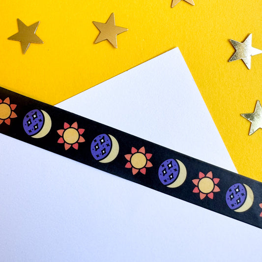 A white piece of paper on a yellow piece of paper with washi tape stuck across the papers. The tape is black with illustrations of suns and moons on it. 