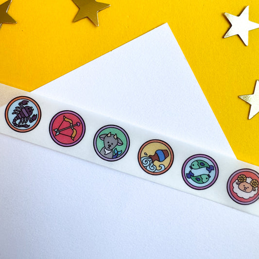 A white piece of paper on a yellow pice of paper with a piece of washi tape running over it. The tape has a white background with circles of illustrations for each zodiac sign along it.