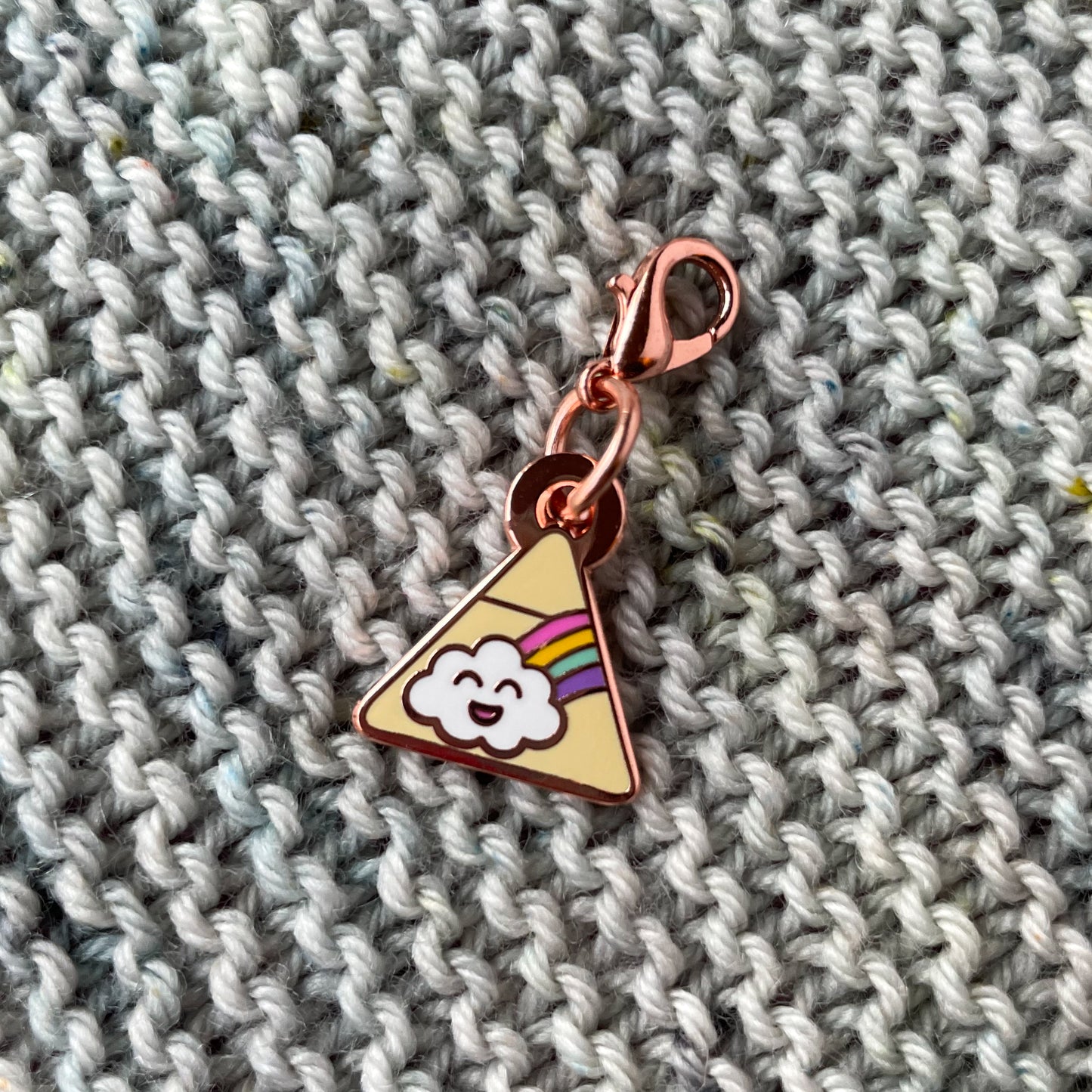 A triangle charm with a happy rainbow cloud on it. The charm is on a background of grey garter stitch knitting.
