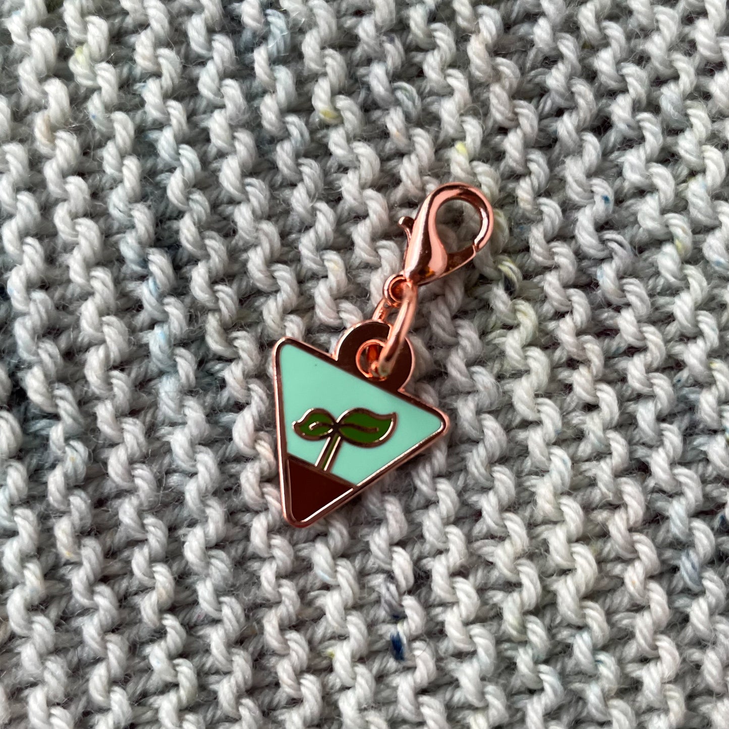 An upside down triangle charm with a small plant sprout on it, sitting on a background of hand knit garter stitch fabric in grey yarn.