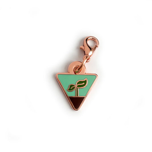 A charm in the shape of an upside down triangle with a small plant sprout growing out of some dirt on it. 