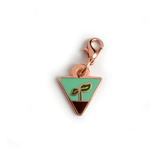 A charm in the shape of an upside down triangle with a small plant sprout growing out of some dirt on it.