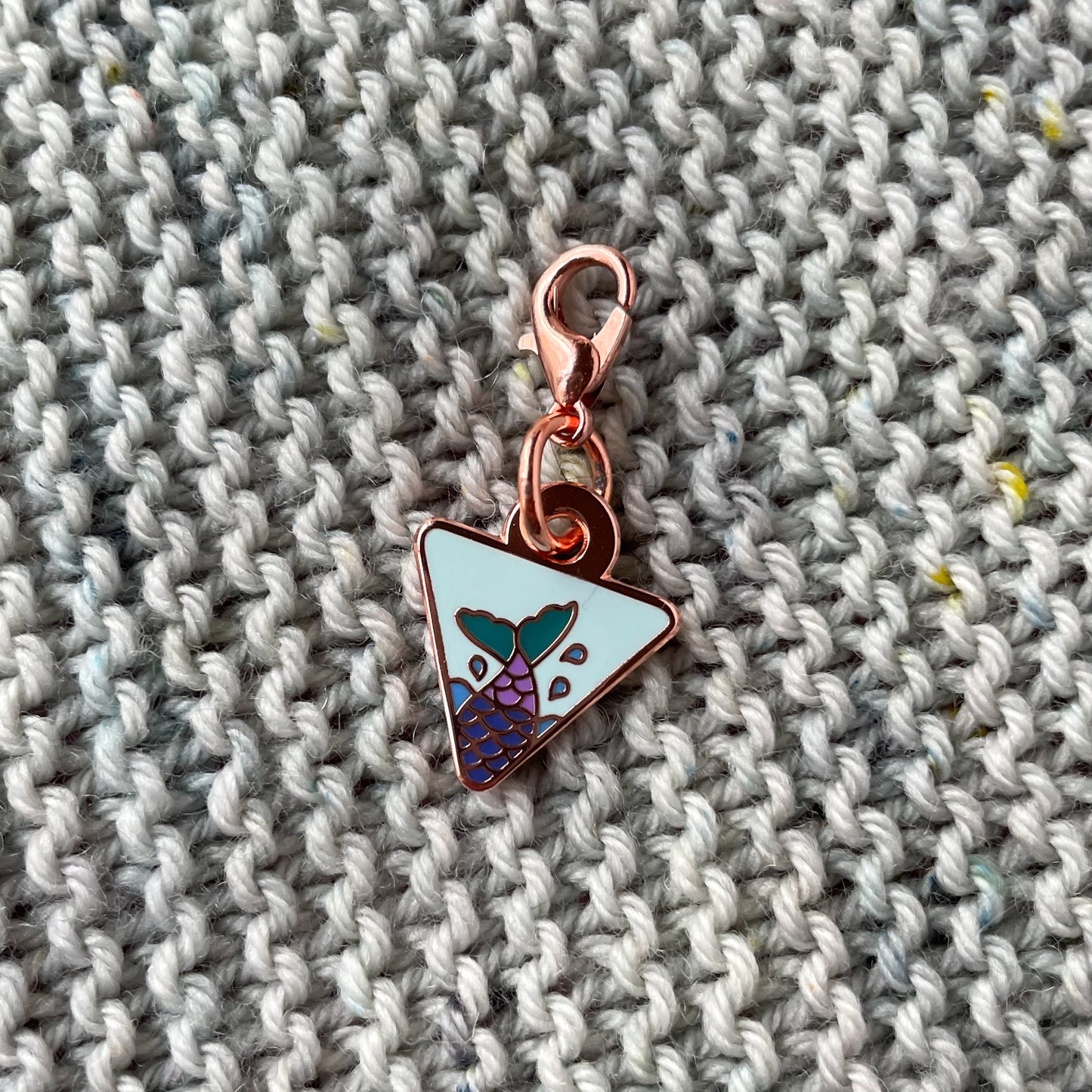 An upside down triangle charm with a mermaid tail in it on top of hand knit garter stitch fabric 