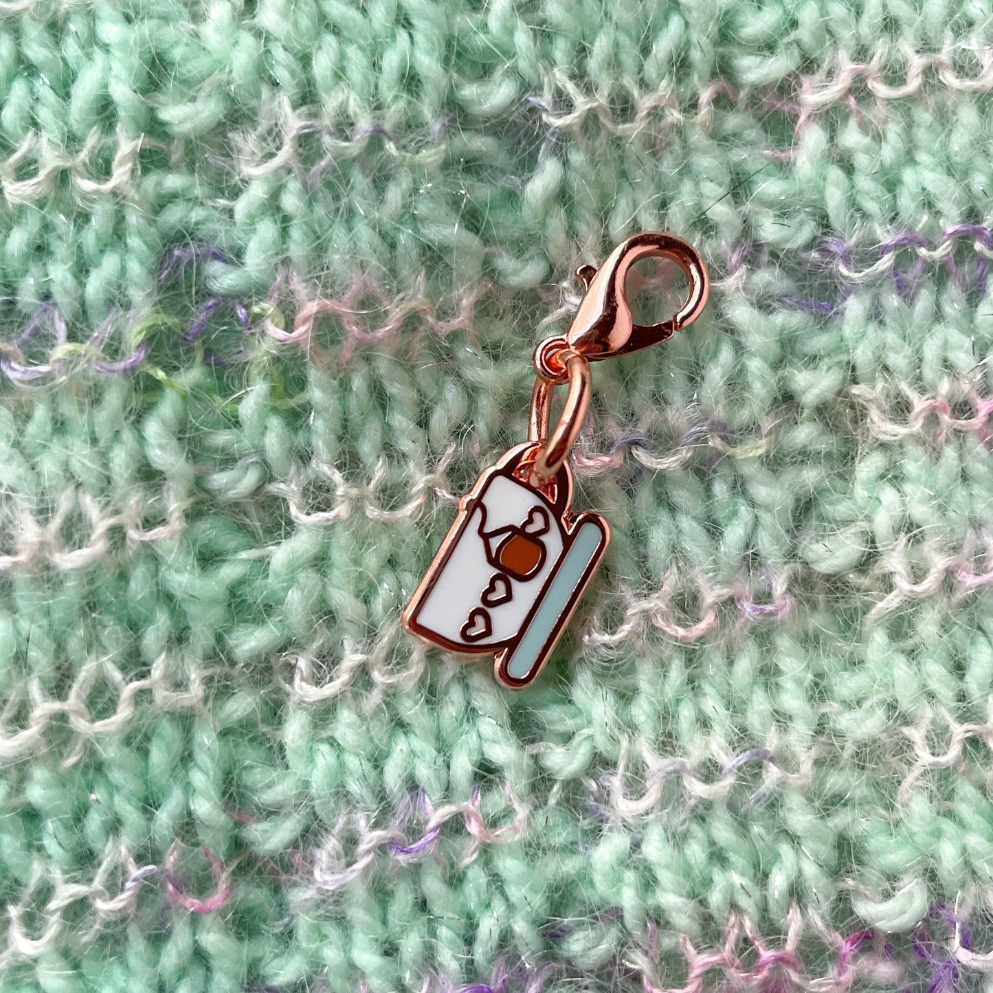 A charm on a lobster claw clasp that is shaped like a teacup with pink hearts on it and a teabag. The charm is on a hand knit background.