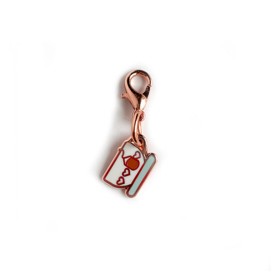 A charm on a lobster claw clasp that is shaped like a teacup with pink hearts on it and a teabag.