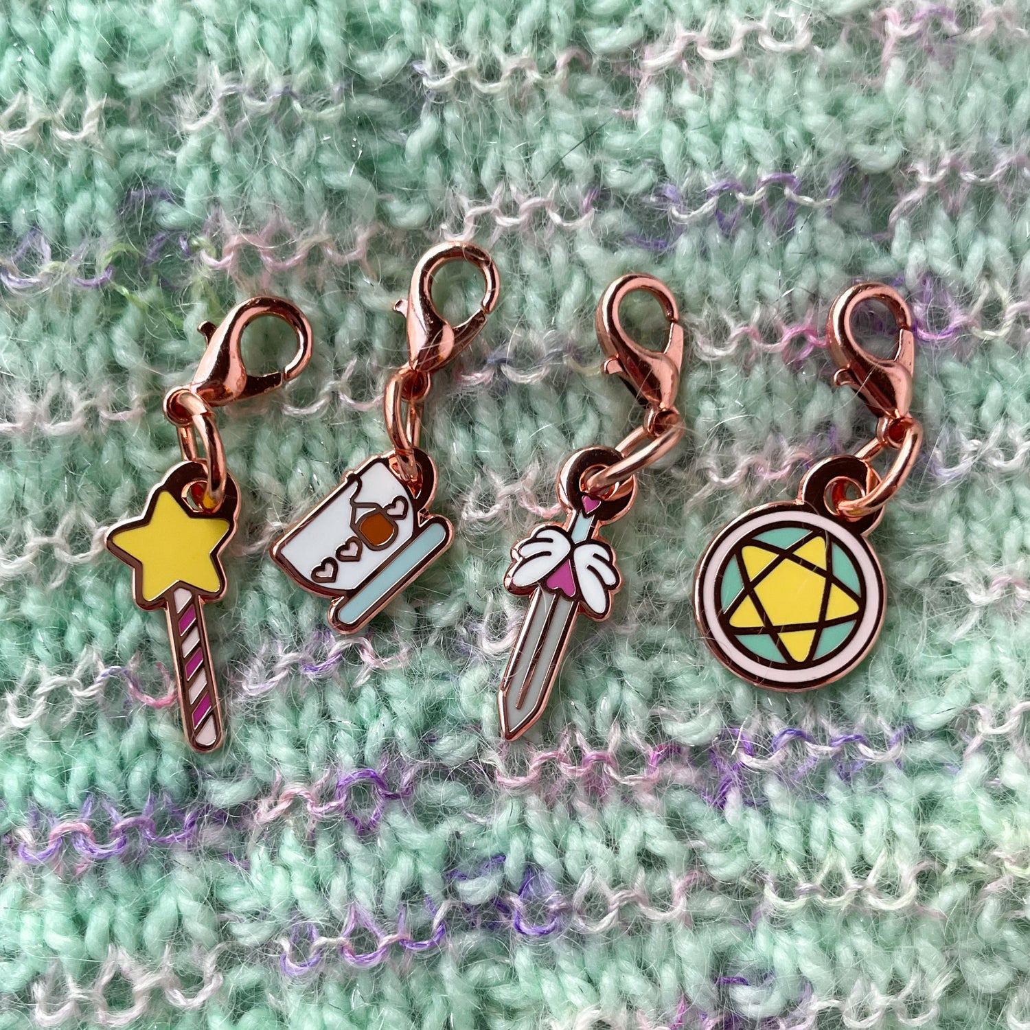 A collection of charms on a hand knit  background. The charms are shaped like a sword, a teacup, a wand, and a pentacle circle.