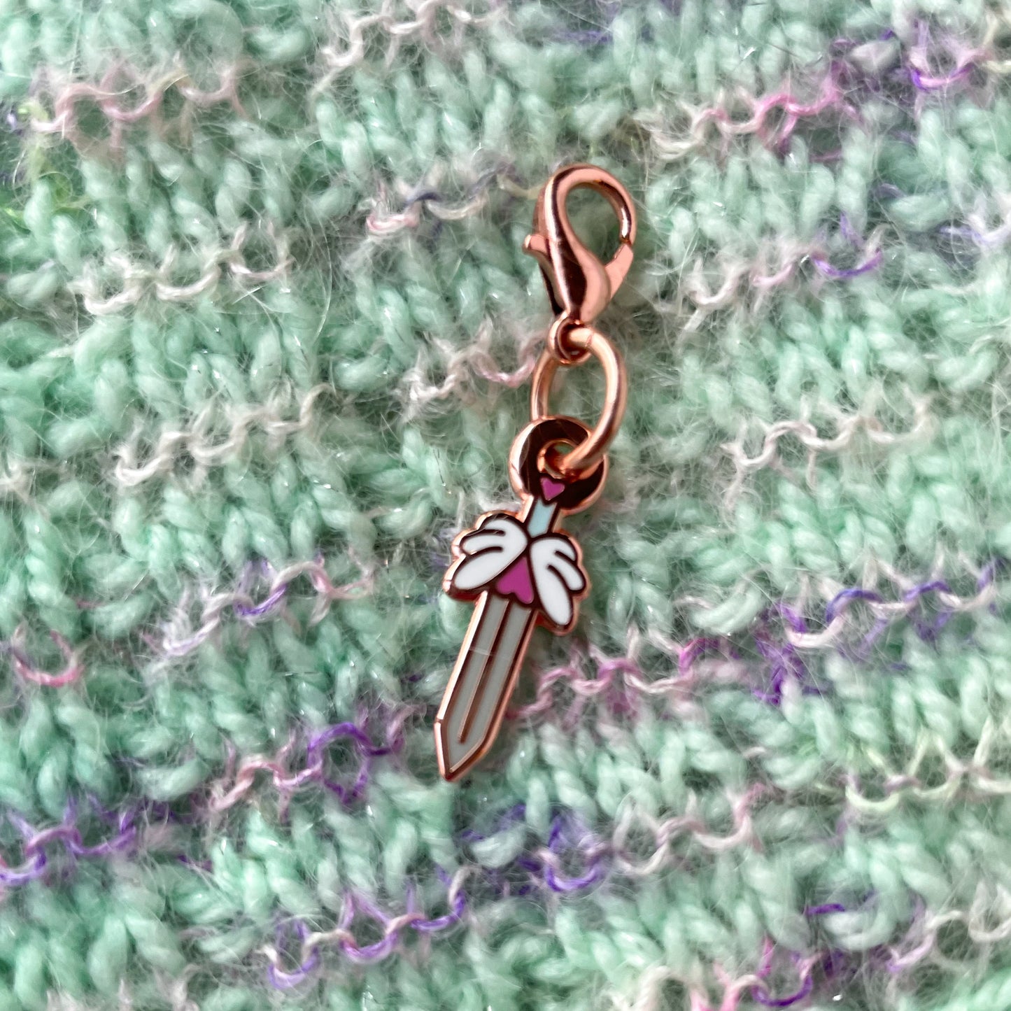 A sword charm on a background of handknit fabric. The fabric is mint and sparkly with stripes of fuzzy pink.
