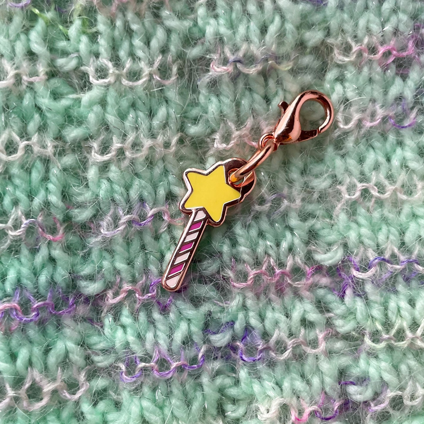 A star wand charm on a background of hand knit fabric. The fabric is mint green with fuzzy pink stripes.