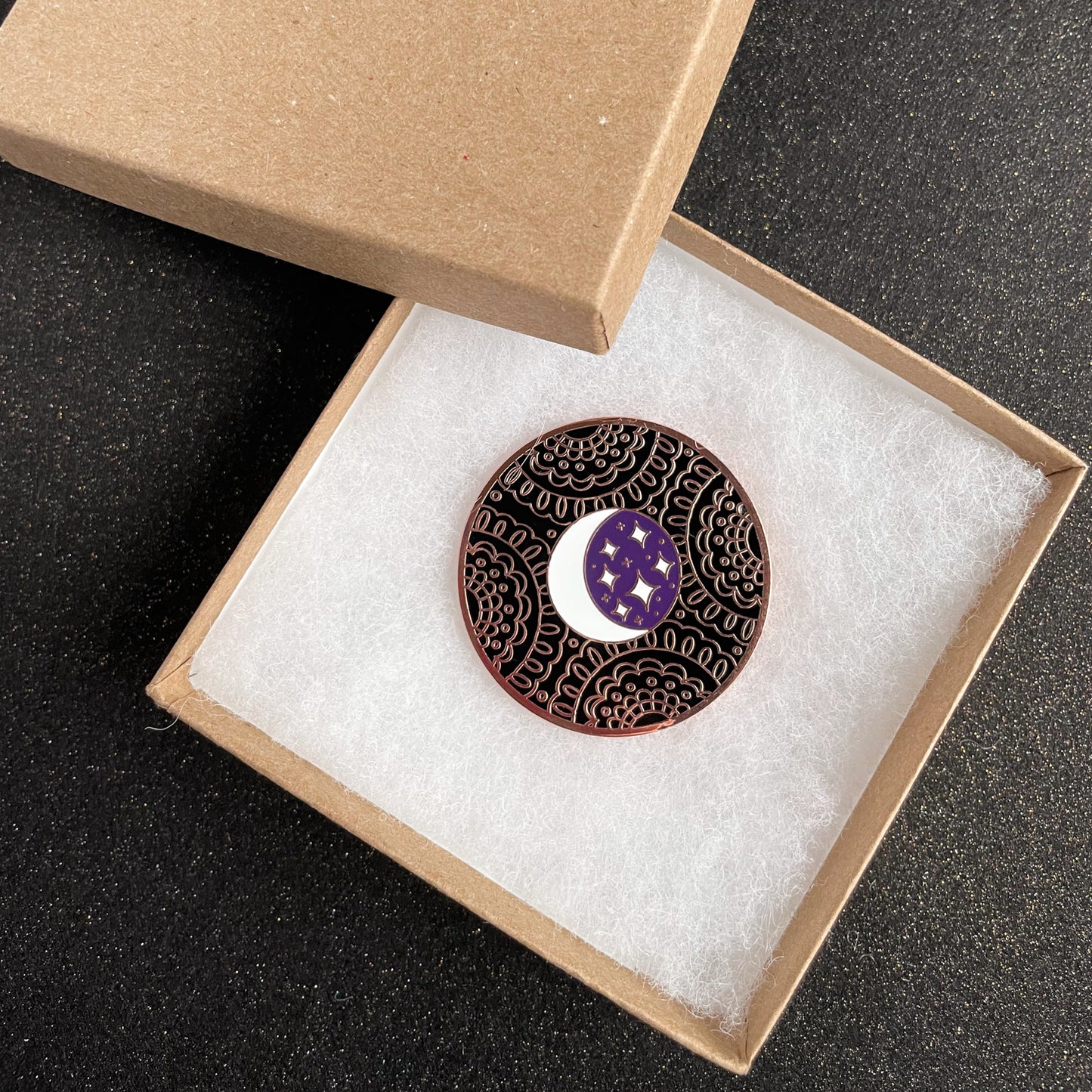 A coin with a moon and stars on it with a black lace background. It is in a kraft paper box on a black glitter background.
