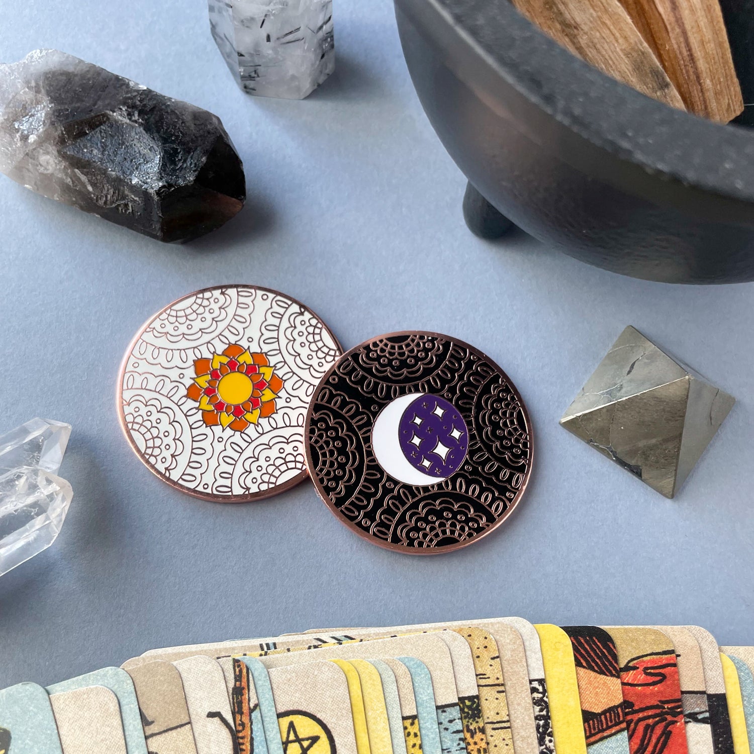 A coin with a sun on a white lace background resting on a coin with a moon and stars on a black lace background. The coins are surrounded by crystals and tarot cards.