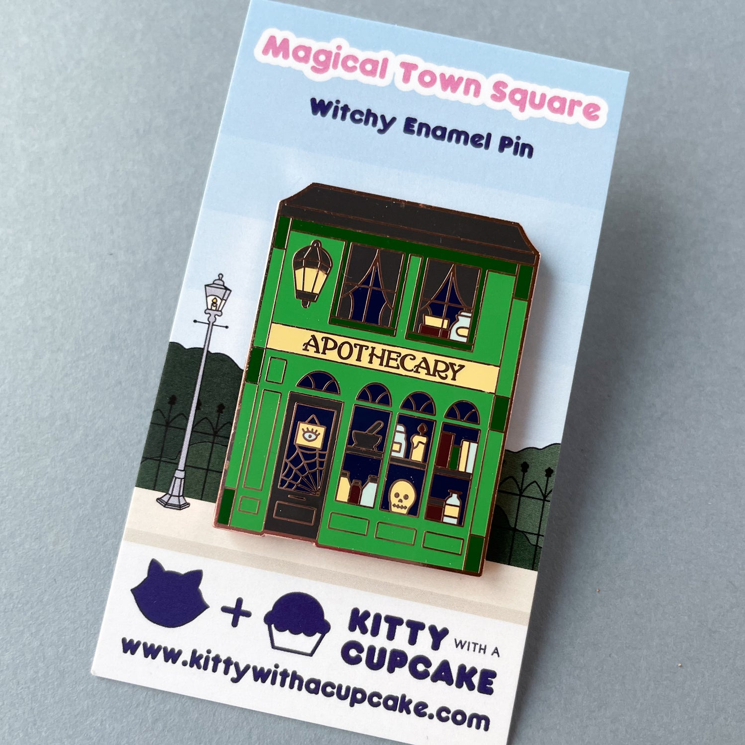 An enamel pin shaped like an Apothecary storefront on a backing card that says "Magical Town Square Witchy Enamel Pin".