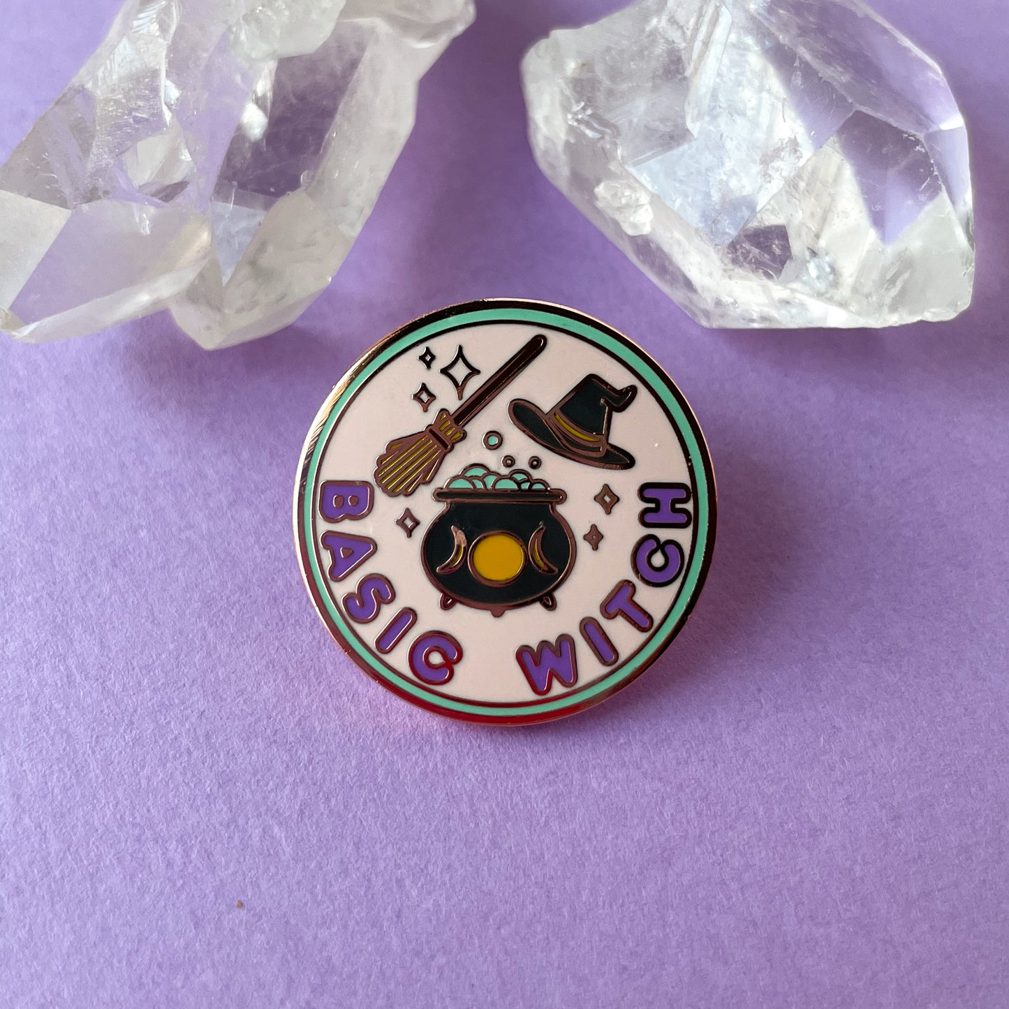A circular hard enamel pin with rose gold metal. There is a mint green border around a pastel pink circle in the background. The bottom of the pin has bubble letters in purple that read "Basic Witch" above this there is a black cauldron with yellow moon phases full of bubbles. There is a broom above this and a black pointed hat to the right of the broom. Sparkles are around all of the objects. This pin is on a purple paper background with some quartz crystals in the background