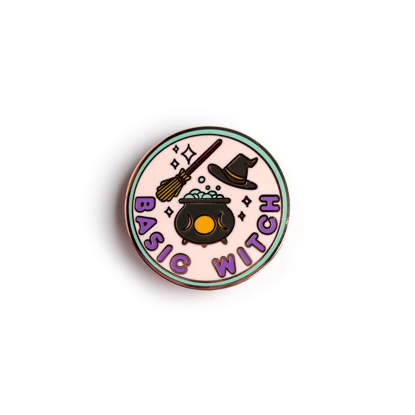 A circular hard enamel pin with rose gold metal. There is a mint green border around a pastel pink circle in the background. The bottom of the pin has bubble letters in purple that read "Basic Witch" above this there is a black cauldron with yellow moon phases full of bubbles. There is a broom above this and a black pointed hat to the right of the broom. Sparkles are around all of the objects. 