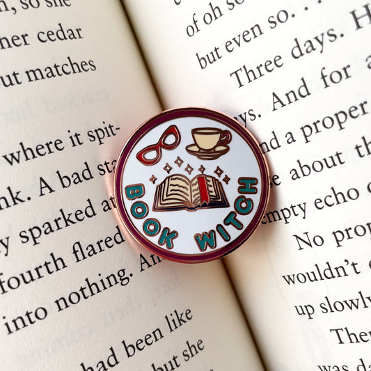 A circular enamel pin sitting in the middle of an open book. The pin says "Book Witch" and has an open book with a bookmark, sparkles, glasses and a teacup on it. 