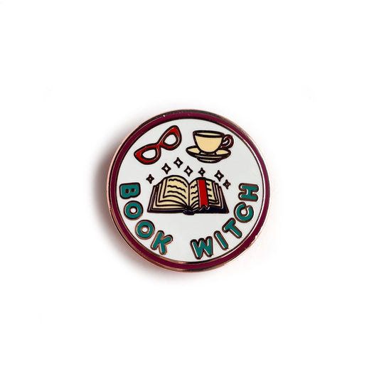 A circular enamel pin with copper metal details. It has a maroon border with a light blue background and turquoise words that say "Book Witch". Also depicted are an open book with a bookmark, a red pair of glasses and a teacup. There are sparkles above the book. 