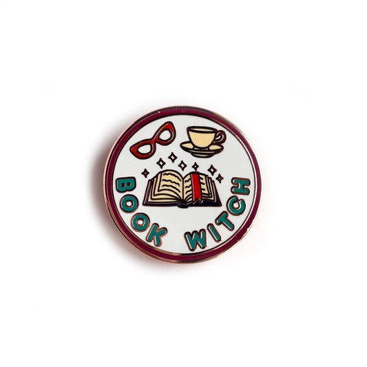 A circular enamel pin with copper metal details. It has a maroon border with a light blue background and turquoise words that say "Book Witch". Also depicted are an open book with a bookmark, a red pair of glasses and a teacup. There are sparkles above the book.