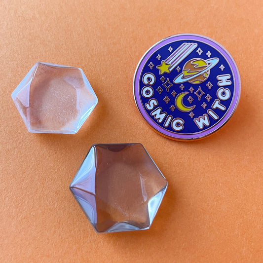 A circular enamel pin on an orange background by two hexagonal quartz crystals. The pin says "Cosmic Witch" and has a moon, shooting star, and the planet Saturn on it.