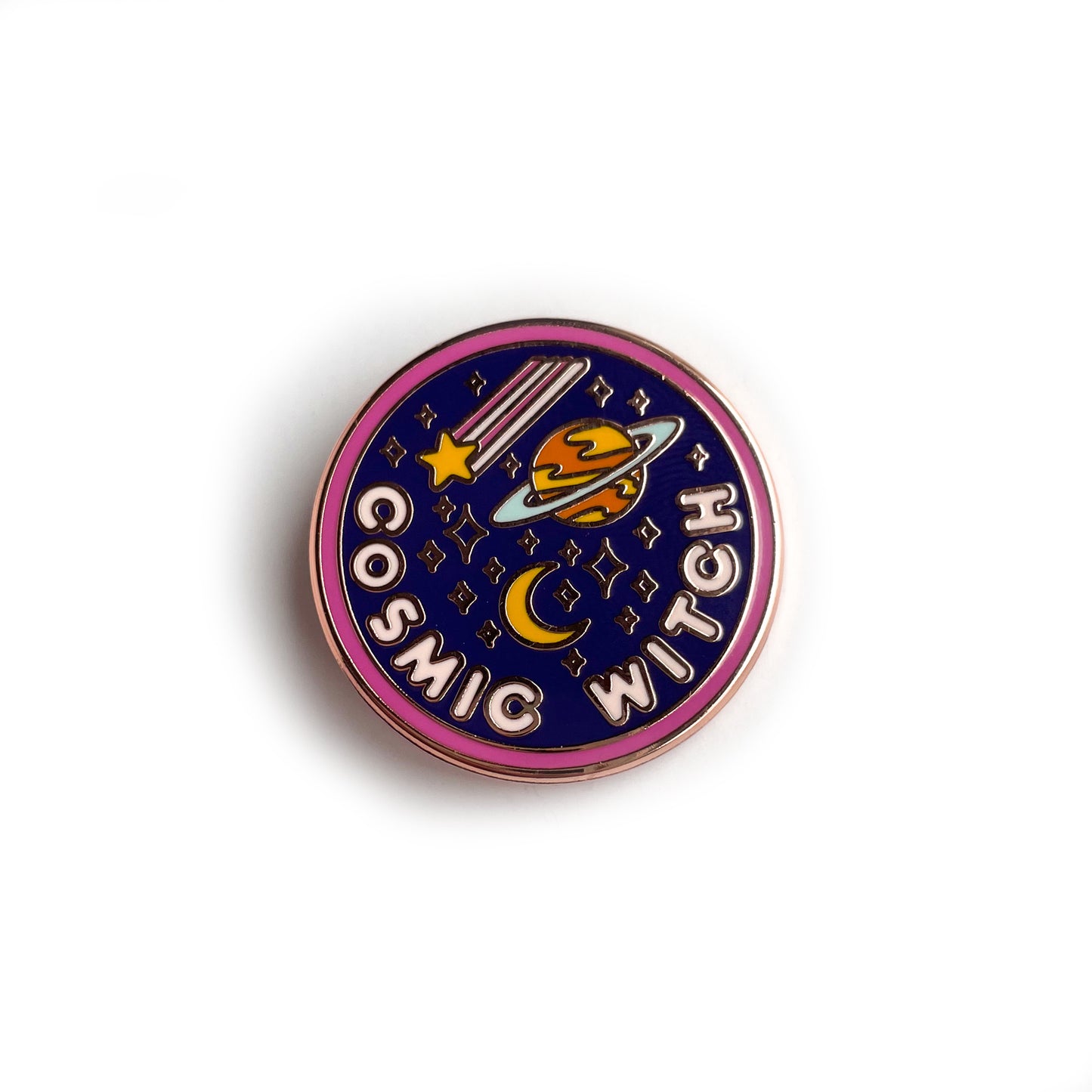 A circular enamel pin. It has a hot pink border, a navy background and light pink letters that read "Cosmic Witch." It also has stars, a crescent moon, a shooting star, and the planet Saturn depicted on it. 