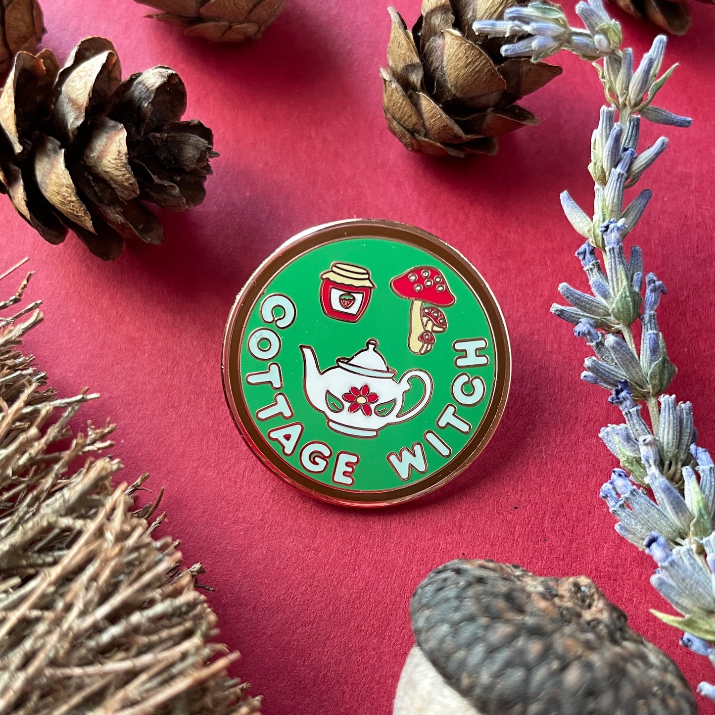 A circular enamel pin on a red background surrounded by small pine cones, an acorn, broom bristles, and a lavender sprig.