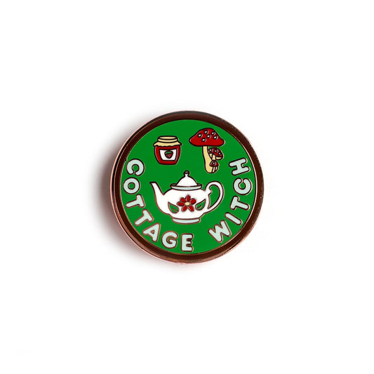 A circular enamel pin. It has a brown border with a sage green background and letters in light blue that read "Cottage Witch". It has a floral teapot, strawberry jam jar, and red mushrooms depicted on it.
