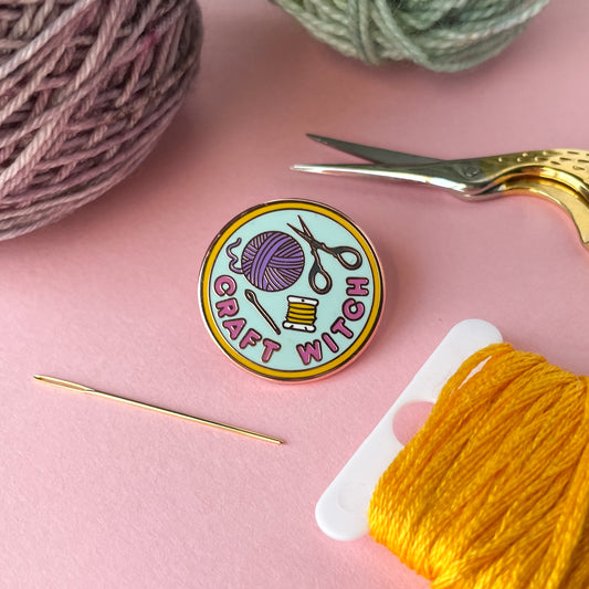 A circular enamel pin on a pink background surrounded by a needle, scissors, embroidery floss and yarn. The pin reads "Craft Witch". 