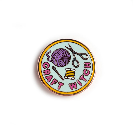 A circular enamel pin. It has a yellow border with a light blue background and hot pink letters that read "Craft Witch". It has a needle, embroidery floss, a purple yarn ball and scissors depicted on it. 