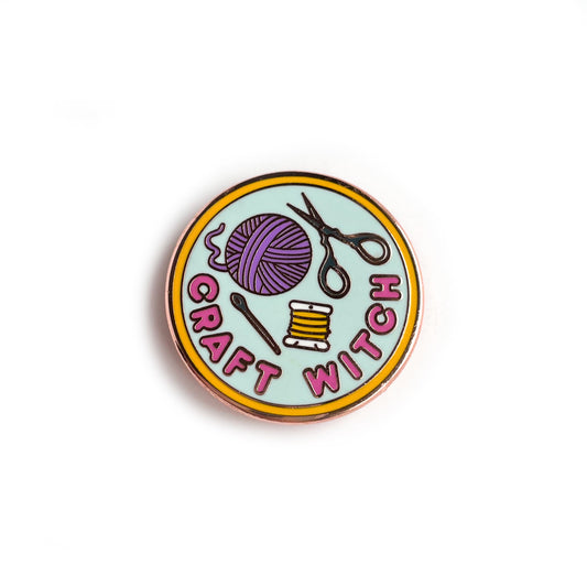 A circular enamel pin. It has a yellow border with a light blue background and hot pink letters that read "Craft Witch". It has a needle, embroidery floss, a purple yarn ball and scissors depicted on it.