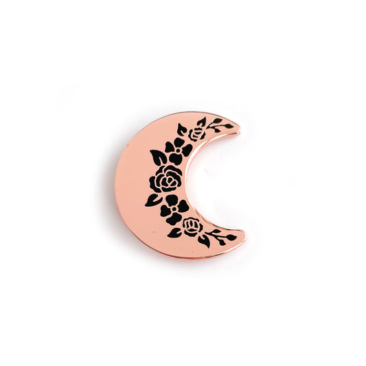 A crescent shaped enamel pin in rose gold metal with black floral shapes along the inside edge.