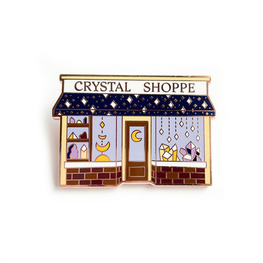 An enamel pin in the shape of shop with the words "Crystal Shoppe" on it. It has a door with a crescent moon on it. The windows are full of crystals in yellow and purple. The awning of the shop has sparkling stars on it and under the front windows are bricks.