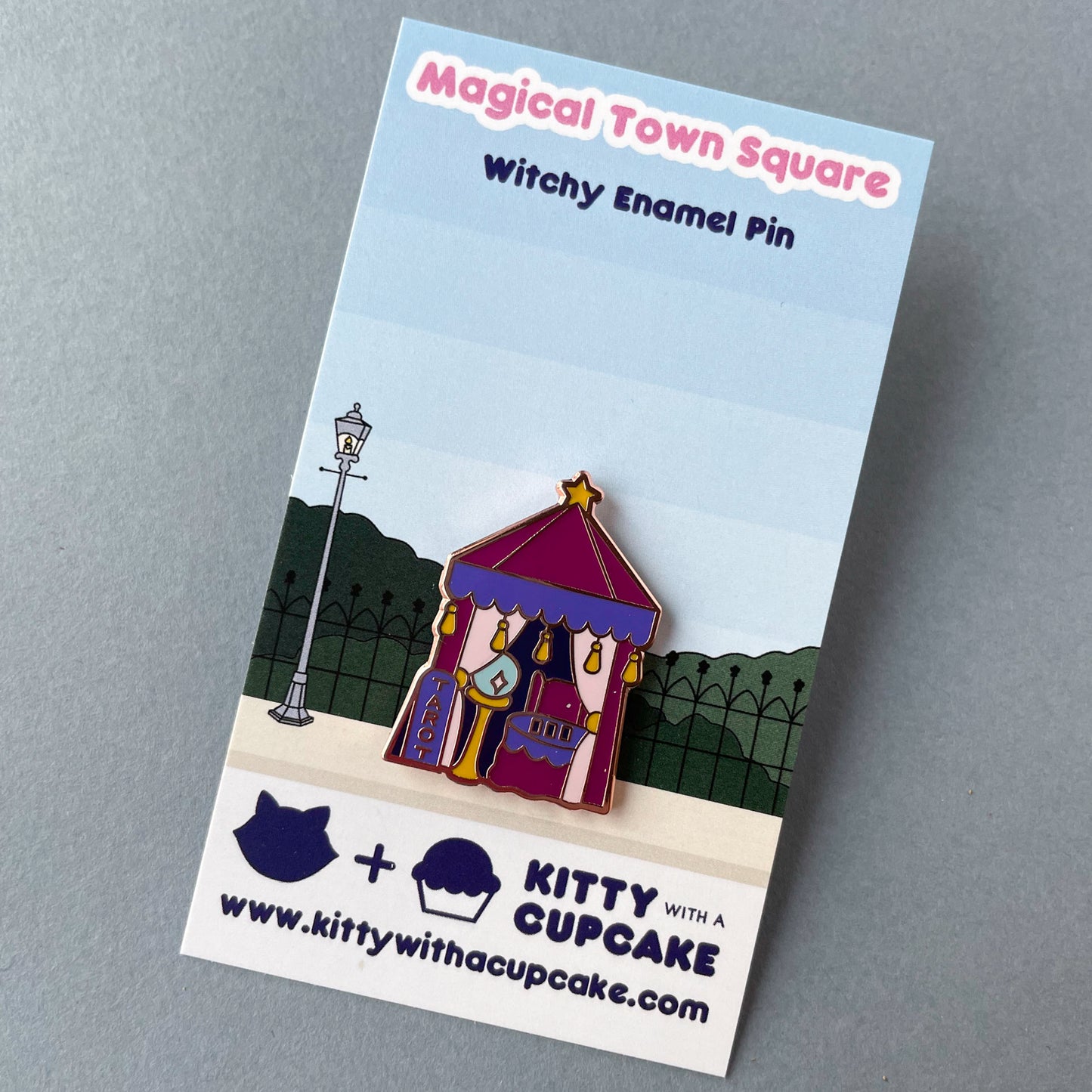 A tarot tent pin on its card packaging. The card reads "Magical Town Square Witchy Enamel Pin" and has a street with a fence and a light post printed on it.