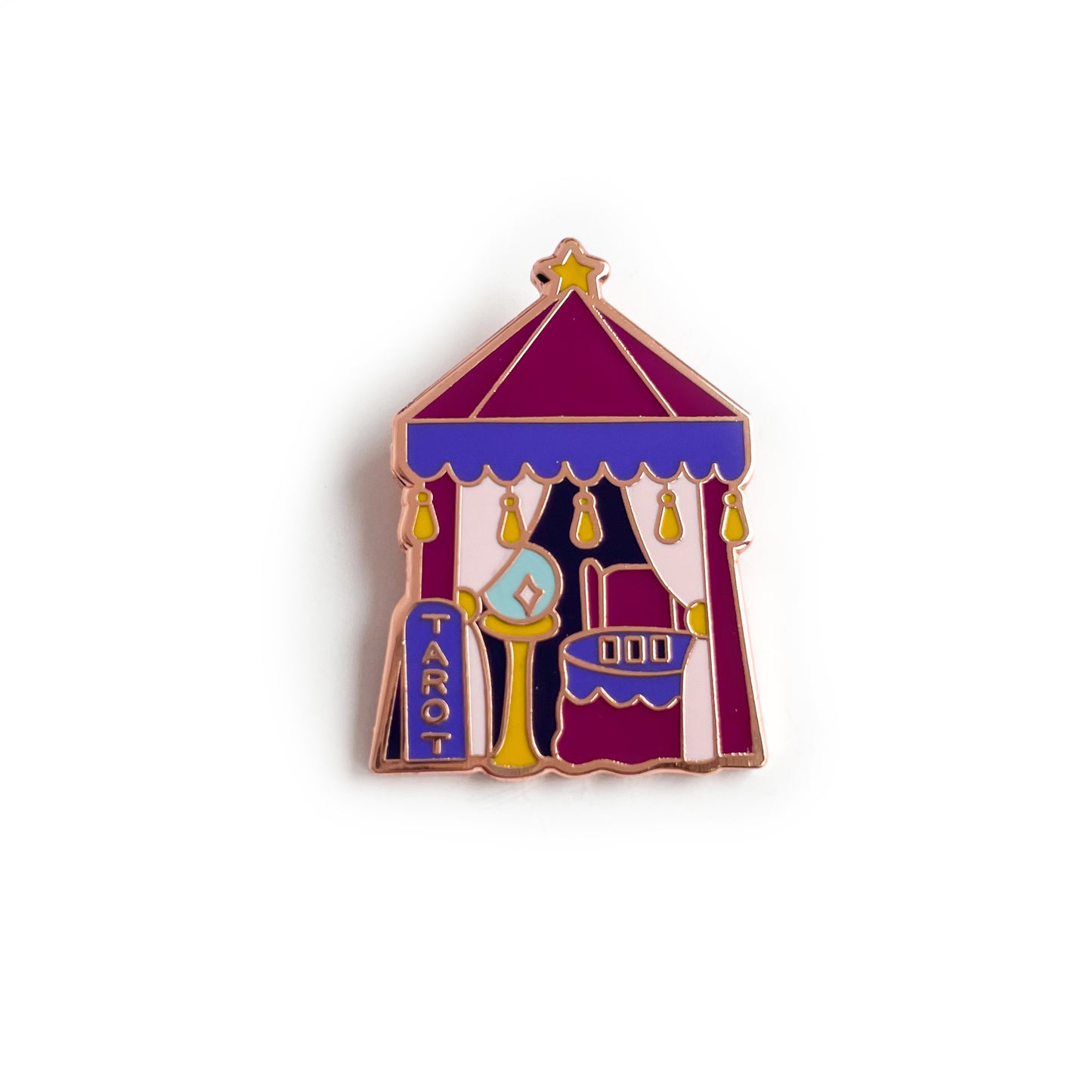 An enamel pin of a small maroon tent. There is a sign outside the tent that reads "Tarot". Inside the tent behind pink curtains are a crystal ball, a chair, and a table with 3 cards laid out.