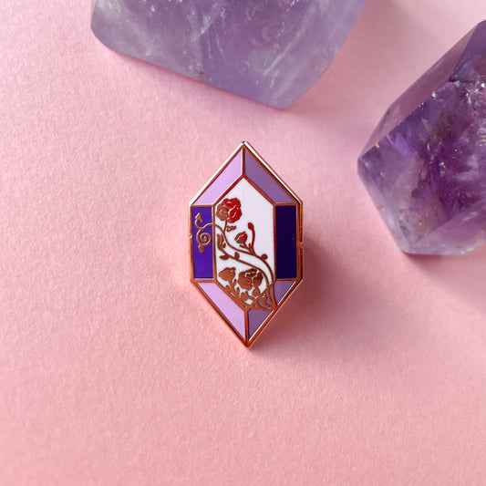 An elongated hexagon shaped enamel pin on a pick background surrounded by amethyst crystals. The pin is various shapes of purple and has floral lines crossing diagonally across it.