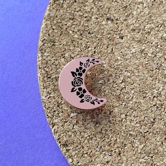 A crescent moon shaped pin in rose gold metal with black floral shapes along the inside edge. The pin is on a corkboard on a purple background.