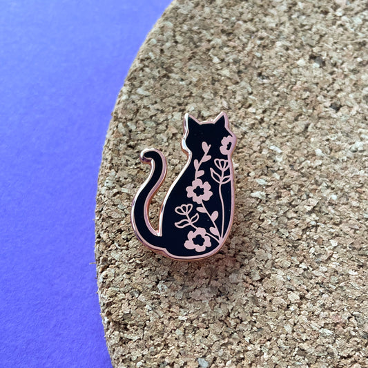 An enamel pin of the back of a black cat with climbing floral vines up its back. The pin is on a cork board on a purple background.