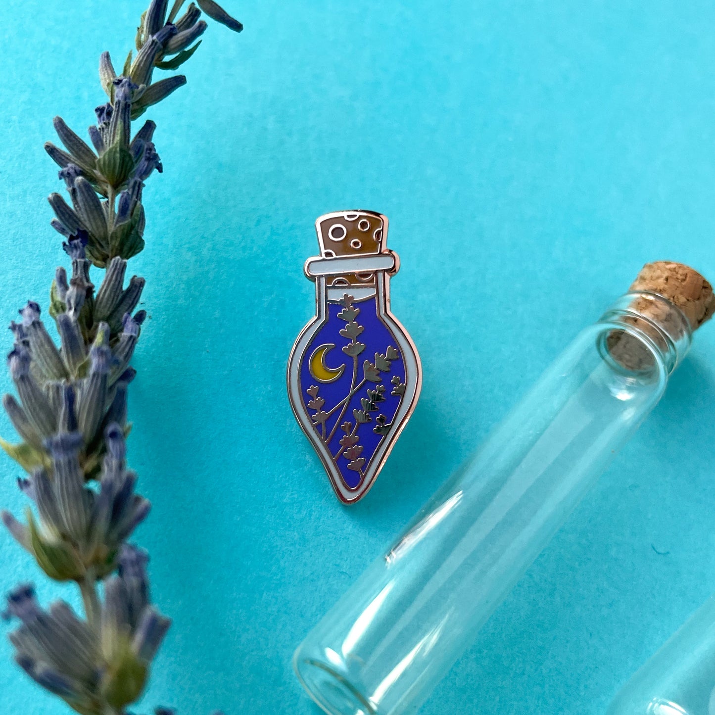 A potion bottle enamel pin on a blue background next to a lavender sprig and a small glass vial.
