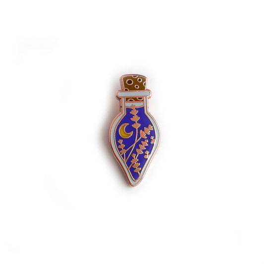 An enamel pin shaped like a teardrop shaped bottle. The bottle has periwinkle liquid withe lavender flowers and a crescent moon in it as well as a cork.