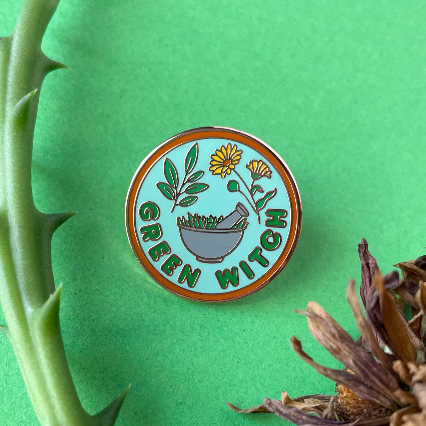 A circular enamel pin on a green background surrounded by dry flowers and a bit of succulent plant. The pin reads "Green Witch" 