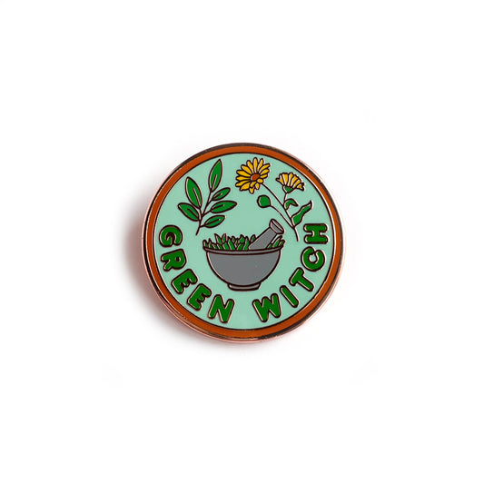 A circular enamel pin with an orange border, mint green background and green letters that read "Green Witch". It has  sage leaves, calendula flowers, and a mortar and pestle depicted on it. 