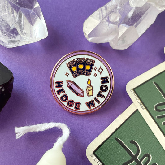 A circular enamel pin on a purple background surrounded by quartz crystals, cards and a candle. The pin reads "Hedge Witch".