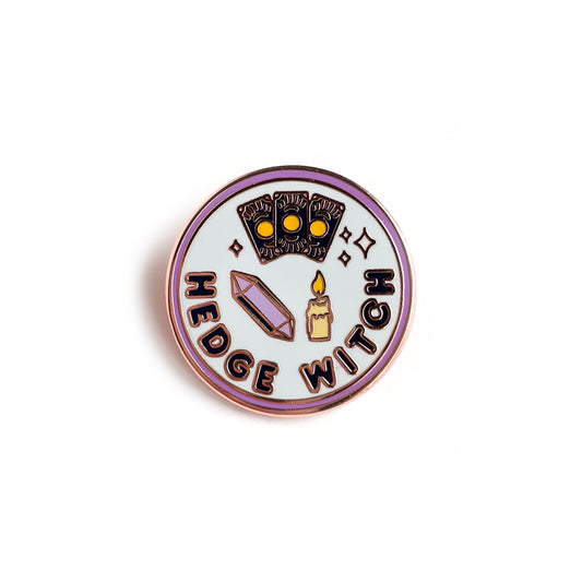 A circular enamel pin with a purple border, light blue background and dark purple words that read "Hedge Witch". There is an amethyst crystal, a candle, and three tarot cards depicted on it.