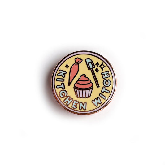A circular enamel pin with a lavender border, pastel yellow background, and words reading "Kitchen Witch". It has a piping bag, cupcake, and spatula depicted on it.