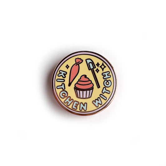 A circular enamel pin with a lavender border, pastel yellow background, and words reading "Kitchen Witch". It has a piping bag, cupcake, and spatula depicted on it.