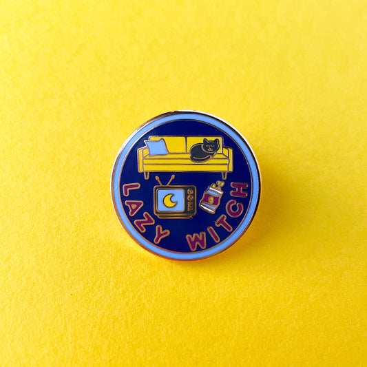 A circular enamel pin on a yellow background. The pin has the words "Lazy Witch" on it.