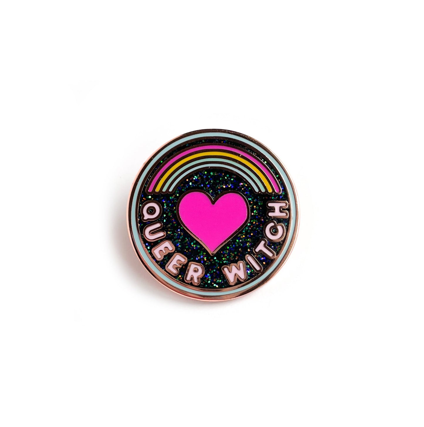 A circular enamel pin with a light blue border, black glitter background and words that read "Queer Witch". It has a rainbow and a heart on it.