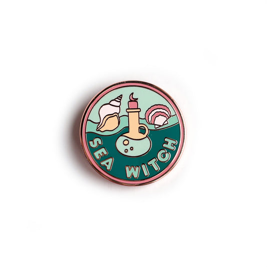 A circular enamel pin with a salmon border and waves in the background with the words "Sea Witch" on it. It has a potion bottle, a scallop and conch shell on it.