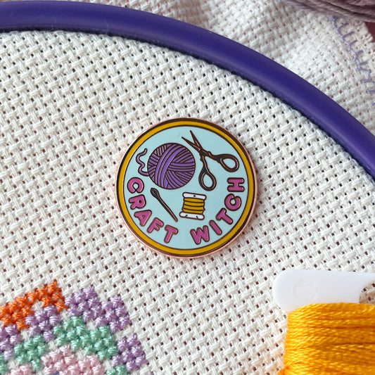 An enamel needle minder shaped like a circle with the words "Craft Witch" and a yarn ball and scissors on it. The item is displayed on an embroidery hoop.
