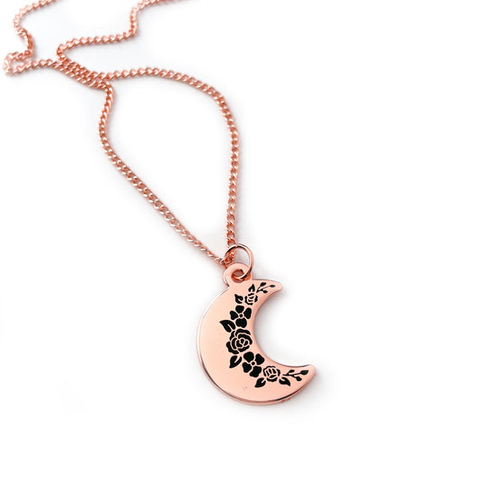 A crescent moon pendanct with black floral silhouettes along the inside edge. It has a copper colored chain. 