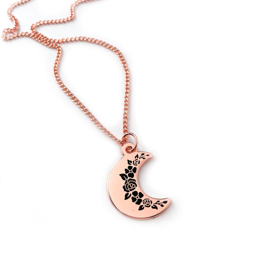 A crescent moon pendanct with black floral silhouettes along the inside edge. It has a copper colored chain.